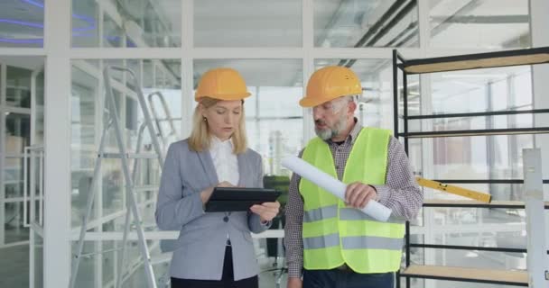 Ritratto di donna biondo esecutivo piuttosto fiducioso-direttore principale del progetto di costruzione ispezionare con ingegnere barbuto anziano o lavori di costruzione appaltatore in cantiere al chiuso — Video Stock