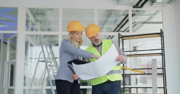 Atractivo gerente principal de mujer rubia altamente calificada y confiada en hardhat discutiendo detalles del proyecto de construcción junto con el ingeniero profesional barbudo mayor al inspeccionar las obras de construcción — Vídeos de Stock