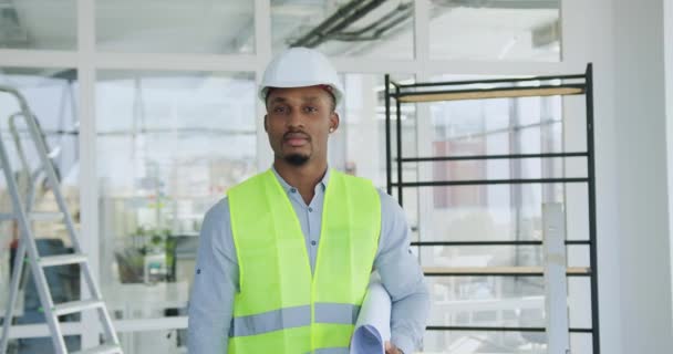 Atractivo ingeniero afroamericano serio confiado en hardhat y chaleco posando en la cámara con plano en el sitio de construcción en el interior — Vídeos de Stock