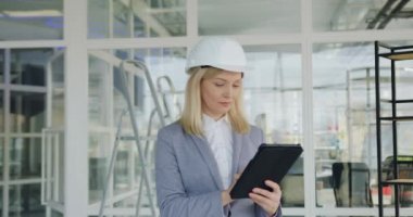 Çekici, kendinden emin, koruyucu kasklı kadın mühendis tamamlanmamış ofis binasında tablet bilgisayar üzerinde çalışıyor ve kameraya bakıyor.