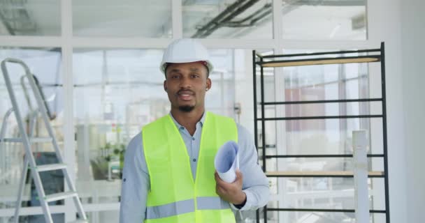 Portrait of likable positive experienced young black-skinned specialist of building company which holding in hands draft with building plan and looking at camera inside in unfinished office building — Stock Video