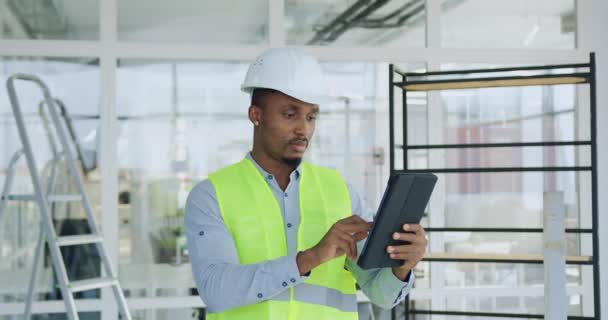 Close-up van aantrekkelijke geconcentreerde professionele zwart-gevilde bouwer in speciale uniforme controle details van het bouwplan op tablet pc bij binnenkomst in het gebouwde kantoorgebouw — Stockvideo