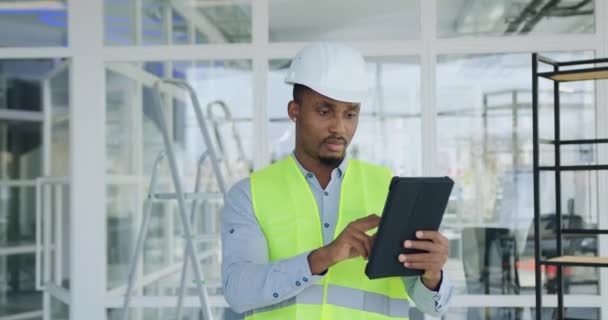 Ritratto di simpatico esperto afroamericano professionista di fiducia laborioso di costruzione di società in uniforme speciale che lavora su tablet pc in un edificio per uffici non finito e guardando la fotocamera — Video Stock