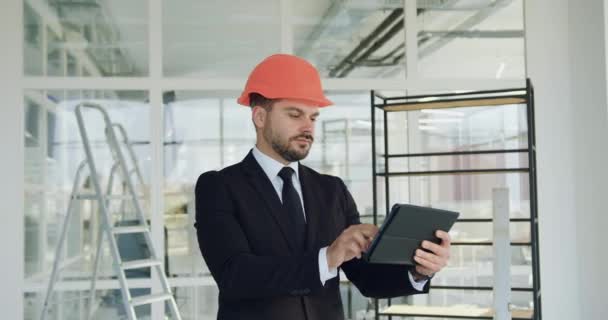 Přední pohled na atraktivní koncentrovaný stylový vousatý manažer stavební firmy v hardhatu, který kontroluje novou kancelářskou budovu a revizní plán na tabletu ks — Stock video
