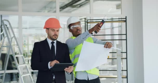 Likable fiducioso esperto manager barbuto e appaltatore afro-americano in caschi in piedi insieme nel bel mezzo del cantiere al chiuso e discutere i dettagli del piano — Video Stock