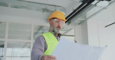 Özel üniformalı çekici, profesyonel sakallı müteahhit tamamlanmamış ofiste planları tutuyor ve tüm detayları plana göre kontrol ediyor.