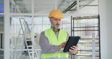 Tamamlanmamış bir binada dururken tablet bilgisayar kullanan iyi görünümlü, kendine güvenen, kendinden emin, kasketli, sakallı ve yelekli yaşlı bir inşaatçının portresi.
