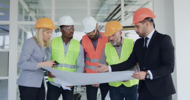 Agradable equipo multirracial experimentado y confiado de especialistas de la empresa de construcción que revisionan el plan de construcción y hablan mientras están de pie en el sitio de construcción en el interior — Vídeos de Stock