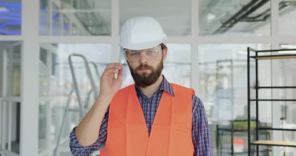 Beau positif confiant constructeur barbu professionnel en uniforme spécial enlever les lunettes de protection et regarder la caméra sur le chantier de construction à l'intérieur — Video