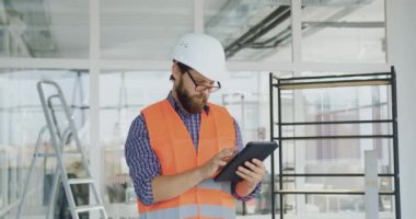 Yakışıklı, sakallı, baret, gözlüksüz ve yelek giyen, yeni inşa edilmiş ofiste tablet bilgisayar kullanan ve kameraya samimi bir gülümsemeyle bakan yetenekli, becerikli bir müteahhit.