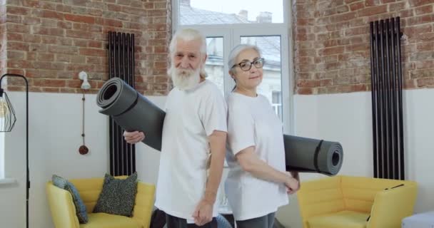 Portrét krásně se usmívající aktivní 60-70. let muž a žena ve sportovním oblečení, které pózují na kameře s rohožemi v obývacím pokoji a ukazují gesto — Stock video
