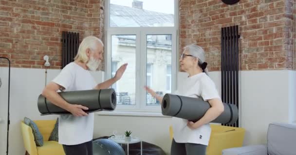 Atractiva y sonriente pareja de ancianos deportistas felices en ropa deportiva dando chocar los cinco antes de empezar a hacer ejercicios en las esteras en casa — Vídeo de stock