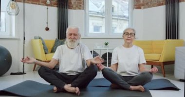 Spor kıyafetleri içinde, evde yoga egzersizleri yaparken gözleri kapalı minderlerin üzerinde meditasyon yapan güzel sakin 60-70 'lerin çifti.