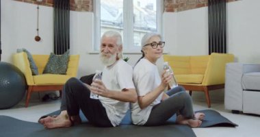 Çekici, kendine güveni olan, sakin, emekli sporcu bir çift paspasın üzerinde oturuyor ve evdeki egzersiz sırasında bir şişe suyla kameraya poz veriyor. Kapatın.