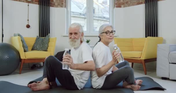 Retrato de hermosa pareja de ancianos deportivos sonrientes en spoerswear que sostiene botellas con agua en las manos mientras se detiene en el entrenamiento conjunto en casa — Vídeos de Stock