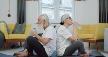 Çağdaş oturma odasındaki paspasların üzerinde dinlenen ve ortak eğitim sırasında şişelerden çıkan tatlı suyun tadını çıkaran sakin, aktif bir spor giyim çifti.