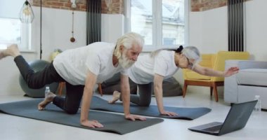Kendine güveni tam, aktif bir emekli çift evde yoga minderleri üzerinde esneme egzersizleri yapıyor ve bilgisayar başında video izleniyor.