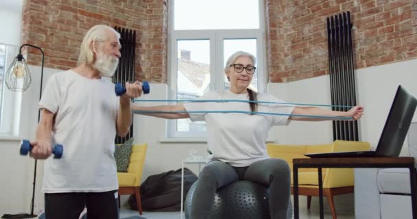 Dobře vypadající pozitivní sportovní starší pár cvičení společně doma s činky a strečink kapela při sledování fitenss cvičení on-line na notebooku — Stock video