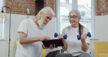 Yakışıklı, gülümseyen sakallı, yaşlı adam tablet kullanırken gözlüklü, aktif karısı evde dambıllarla egzersiz yapıyor.