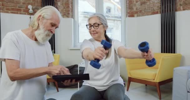 Pretty happy satisfied sporty retired woman in sportswear doing exercises with dumbbells near her bearded husband which looking for something on tablet device — Stock Video