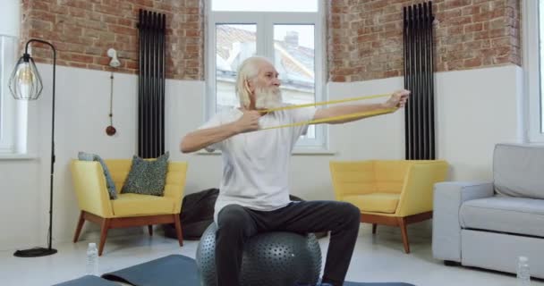 Schöner, konzentrierter, reifer, bärtiger Mann in Sportbekleidung, der auf Fitnessball sitzt und die Hände mit Gummiband dehnt, während er allein zu Hause trainiert — Stockvideo