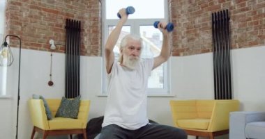 Spor üniformalı, spor ayakkabılı ve evde dambıllarla egzersiz yapan hoş, yoğun, yaşlı, sakallı bir adamın ön görüntüsü.