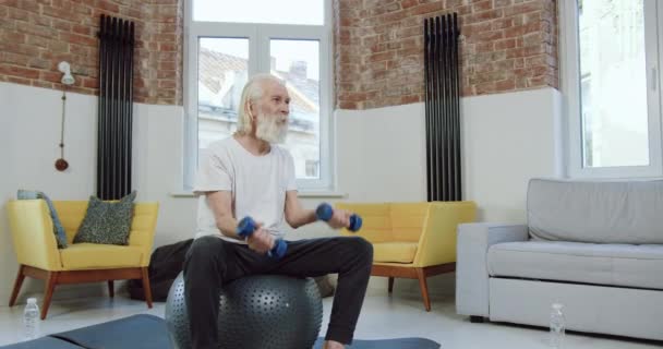 Πορτρέτο της ελκυστική ηρεμία ενεργό ηλικιωμένος γενειοφόρος άνδρας στα αθλητικά που κάθεται σε μπάλα γυμναστικής και κάνει ασκήσεις με αλτήρες κατά τη διάρκεια της κατ 'οίκον προπόνηση — Αρχείο Βίντεο