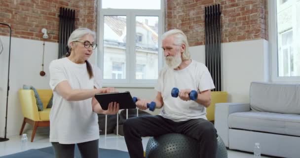 Adorable pareja madura activa positiva en ropa deportiva haciendo ejercicios con pesas simultáneamente viendo lecciones en línea en el dispositivo de la tableta — Vídeos de Stock