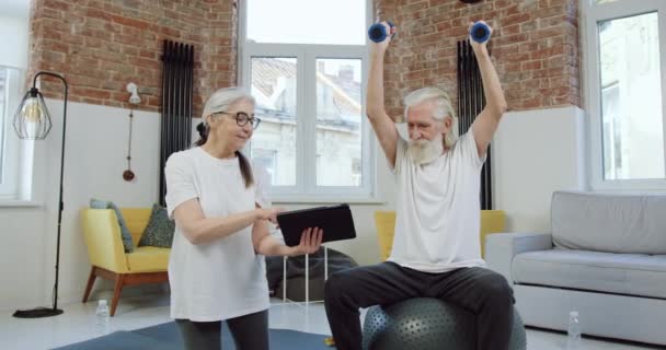 Bonito sorriso ativo barbudo velho fazendo exercícios com halteres e repetindo exercícios que sua esposa positiva mostrando no dispositivo tablet, conceito de treino em casa — Vídeo de Stock