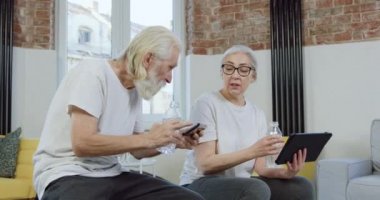 Çekici, gülümseyen, mutlu, yaşlı bir çift, boş vakitlerin tadını çıkarıyor ve oturma odasında yan yana oturup tablet ve telefondaki uygulamaları izliyorlar.