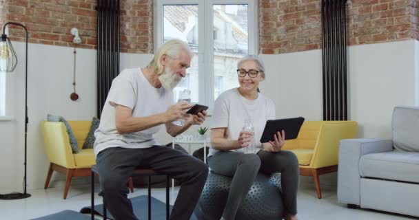 Amar sonriente pareja de ancianos disfrutando de revisiones de aplicaciones de teléfono y tableta mientras se relaja después de ejercicios físicos en casa — Vídeo de stock