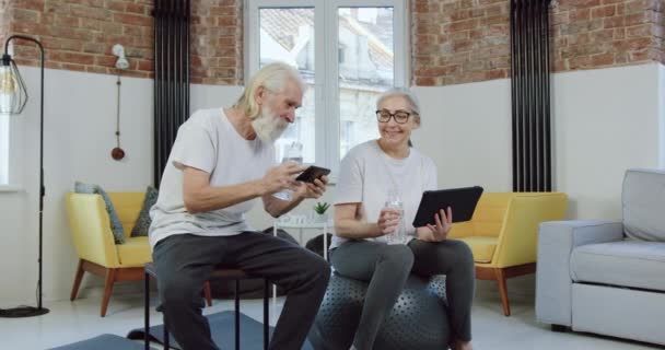 Adorando sorrir casal sênior apreciando revisões aplicativos de telefone e tablet enquanto relaxa após exercícios físicos em casa — Vídeo de Stock