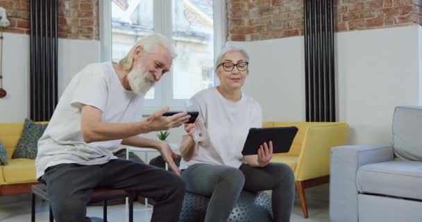 Portrait of adorable cheerful loving retired couple in sportswear which enjoying of revision of funny videos on smartphone during joint leisure at home — Stock Video