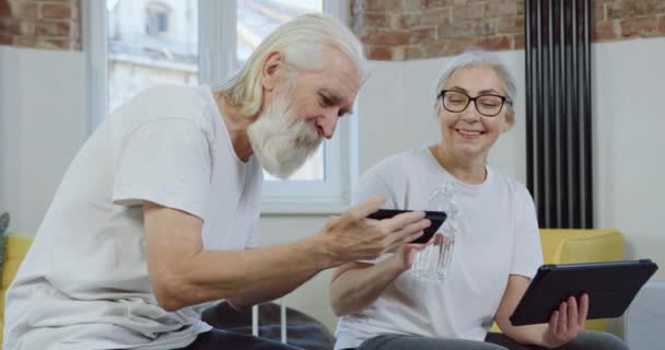 Attractive smiling happy mature couple in white t-shirts having fun together while revisioning funny videos on mobile in beautifully decorated flat — Stock Video