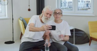Güzel görünümlü, mutlu, spor giyimli, birbirine yakın oturan ve şişeleri eklem egzersizinden sonra suyla tokuştururken selfie çeken çift.