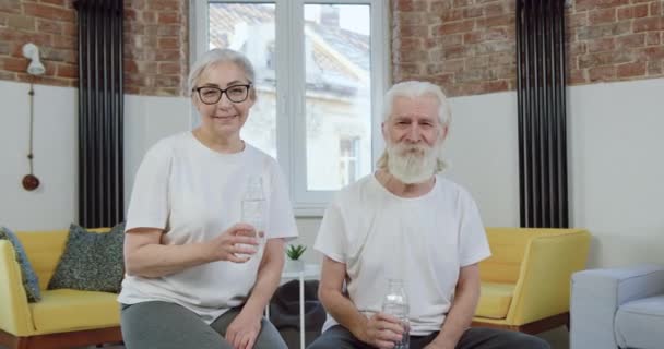 Retrato de atractiva pareja madura activa sin preocupaciones sonrientes que posan en la cámara con botellas de agua durante el entrenamiento conjunto en casa — Vídeos de Stock