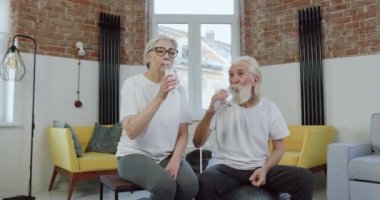 Mutlu, neşeli, aktif, spor üniformalı, su içen yaşlı bir çift. Eklem egzersizinden sonra su içiyor ve iyi bir jest yapıyor.