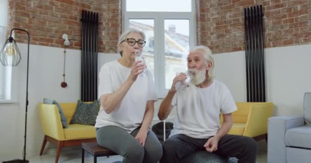 Preciosa feliz alegre activa pareja de edad en uniforme deportivo beber agua después del entrenamiento en casa conjunta y mostrando gesto bien hecho — Vídeos de Stock