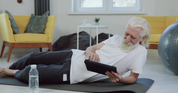 Portret van aantrekkelijke tevreden zorgeloze gepensioneerde man met baard in sportkleding die rust op de mat en gebruikt tablet apparaat om grappige video 's te bekijken — Stockvideo