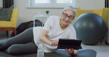 Gülümseyen, mutlu, rahat, gözlüklü, ev elbiseli yaşlı bir kadın evde paspasın üzerinde dinleniyor ve tablet bilgisayarda ilginç videolar izliyor, şişeden su içiyor.