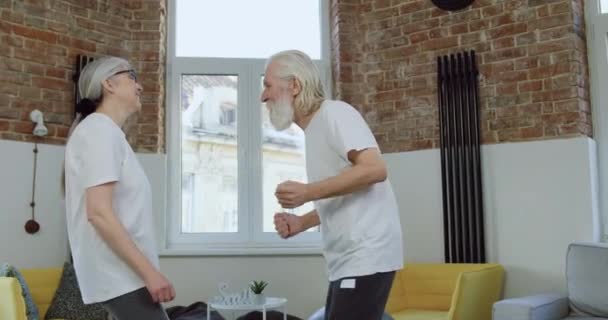 Attractive satisfied funny 70-aged couple in white t-shirts and sports pants dancing under disco music in contemporary living-room while spending weekend at home — Stock Video
