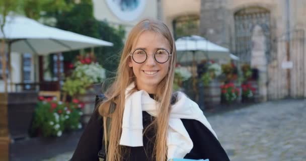 Charmante glimlachende vrolijke moderne jonge vrouw in stijlvolle kleren voor de camera staan tijdens het wandelen op straat met gezellige cafe op zonnige dag — Stockvideo