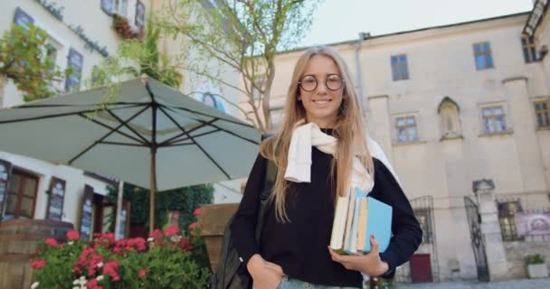 Estudante moderna alegre atraente com livros e mochila posando na câmera perto de belo café de rua na estrada para a universidade — Vídeo de Stock