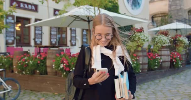 Přední pohled na krásně se usmívající šťastná mladá žena v mladistvých šatech v brýlích, které pomocí telefonu, zatímco stojí v blízkosti letní venku kavárna s krásně zdobené květinami — Stock video