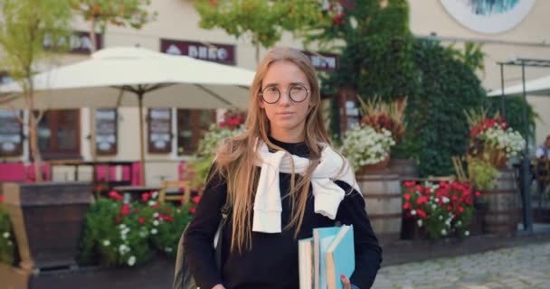 Портрет досить позитивної молодої блондинки студентки в офіційному одязі в окулярах з книгами, які дивляться на камеру на фоні затишного вуличного кафе в сонячний день — стокове відео
