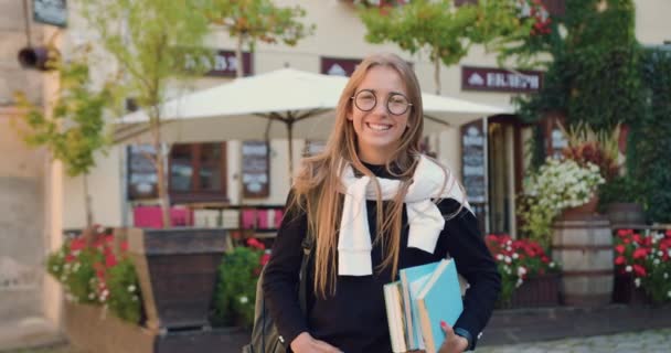 Attraente euforico sorridente giovane ragazza del college bionda in abiti casual in occhiali alla moda tenendo libri in mano e guardando la fotocamera sullo sfondo di caffè all'aperto sulla strada per studiare, da vicino — Video Stock
