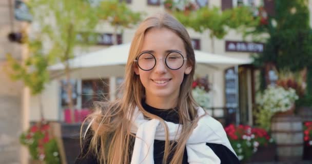 Beautiful picture of charming smiling happy girl in stylish clothes and glasses which standing near cozy street cafe on sunny day and the wind blowing her light long hair — Stock Video