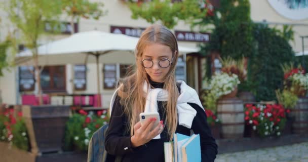 Güzel, gülümseyen, açık renk saçlı, resmi giysiler içinde, elinde kitaplarla gezen ve güneşli bir günde, rahat bir sokak kafesinin yanında cep telefonunu kullanan güzel bir kız öğrenci. — Stok video