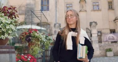 Çekici, mutlu, modaya uygun giysileri olan, elinde kitaplarla cadde kafesinin yanında yürüyen, antik şehir binalarının arka planında çiçekleri olan genç bir kadın.