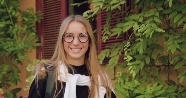 Linda menina sorridente feliz de cabelos claros em óculos elegantes em pé na frente da câmera perto do edifício da cidade antiga com plantas verdes, close-up — Vídeo de Stock